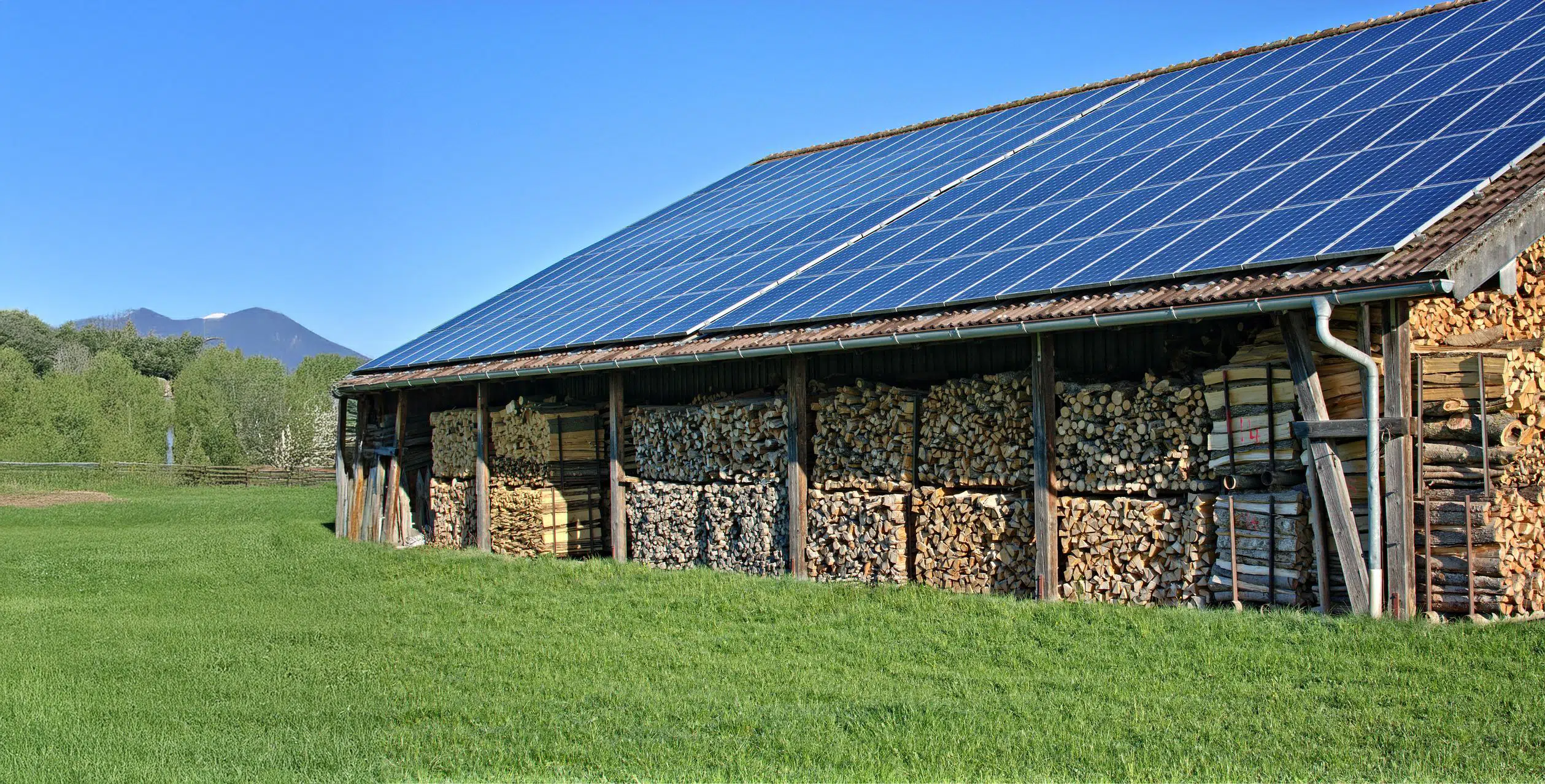 Ein Holzlager unter einem Dach mit Solarpanelen, umgeben von grünem Gras und mit Bergen im Hintergrund.